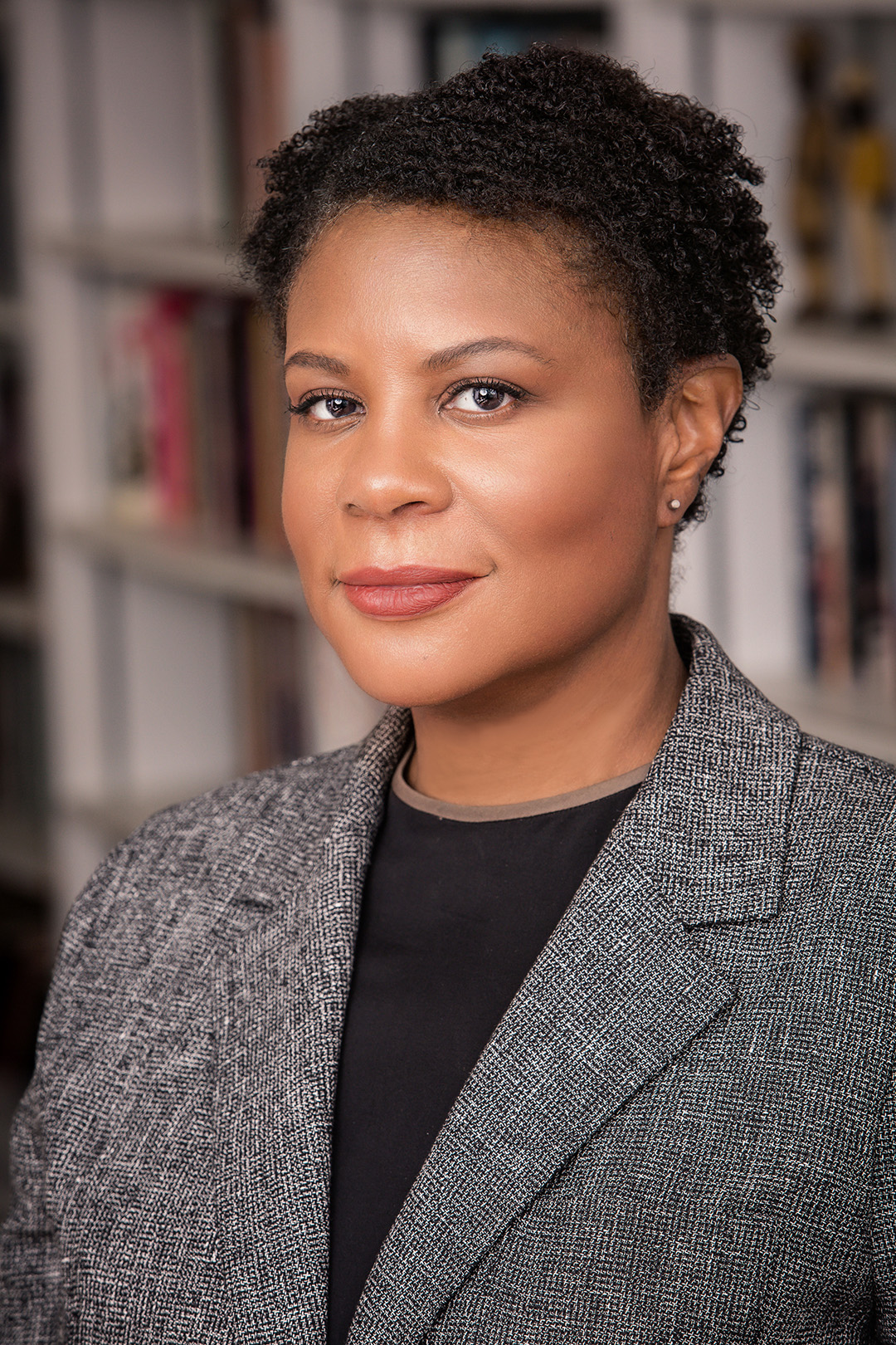 Head and shoulders portrait of an adult with short hair wearing a gray blazer standing in front of a bookshelf